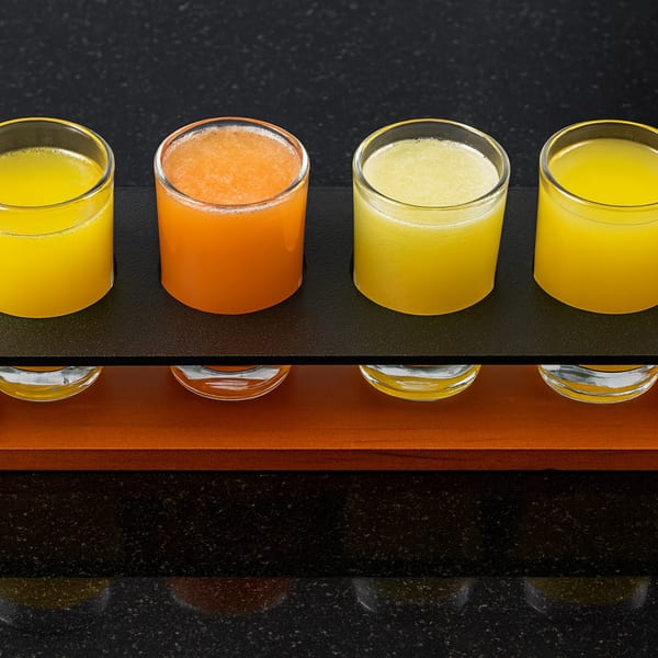 Four glasses of fresh juice, two yellow and two orange, in a dark wooden holder.