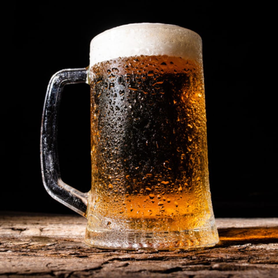 A frosty mug of beer with a foamy head and condensation sits on a rustic wooden table.