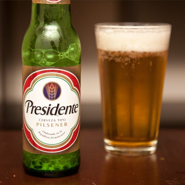 A green Presidente beer bottle next to a glass of foamy beer on a dark table.