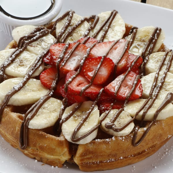 A golden waffle topped with sliced bananas, strawberries, chocolate drizzle, and powdered sugar.