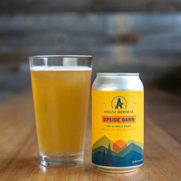 A glass of golden non-alcoholic beer next to an Athletic Brewing Co. Upside Dawn can.